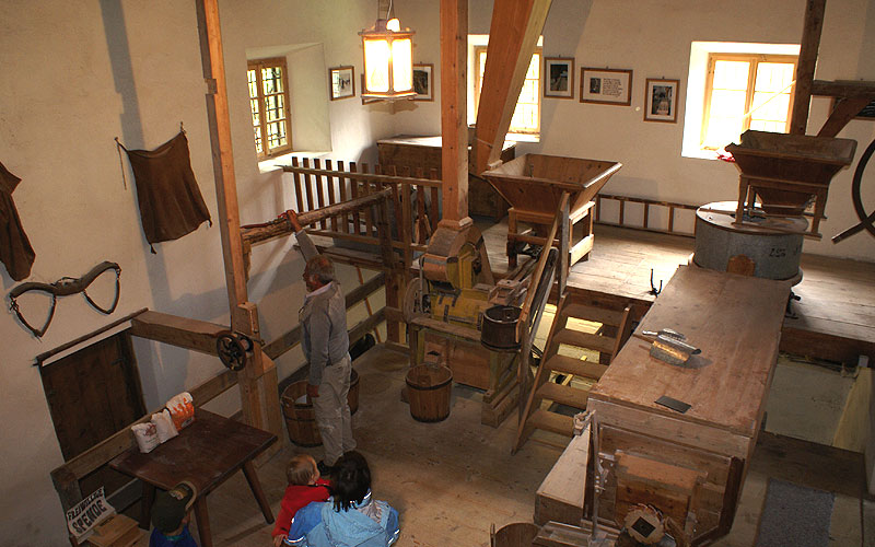 Das Innere einer traditionellen Wassermühle mit hölzernen Maschinen, Eimern, Balken und Besuchern, die die Geräte beobachten. Durch die Fenster fällt natürliches Licht ein.