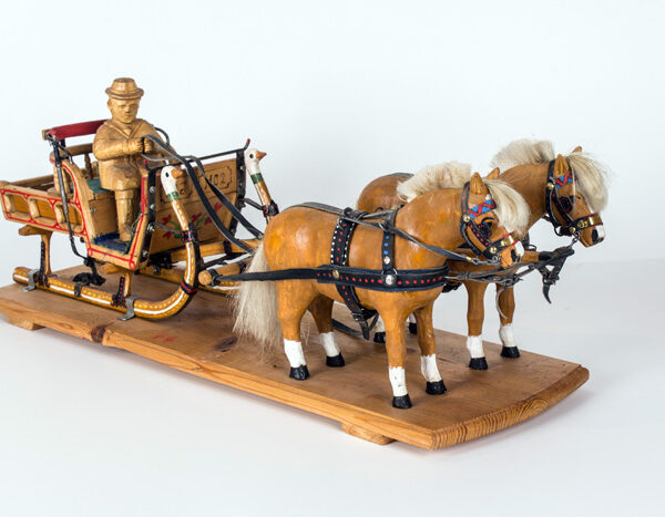 Holzmodell eines Pferdeschlittens mit zwei Pferden und einer Kutscherfigur auf einem Holzsockel vor einem schlichten Hintergrund.