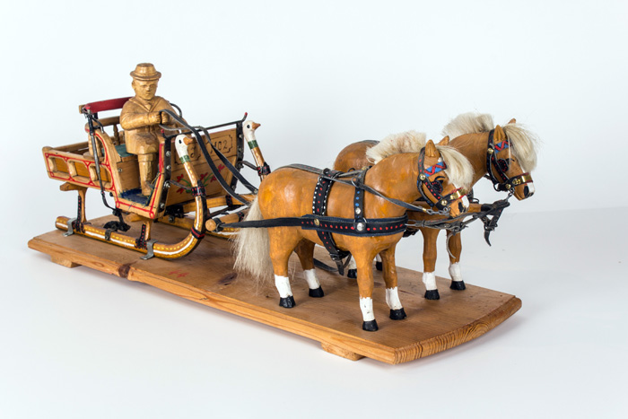 Holzmodell eines Pferdeschlittens mit zwei Pferden und einer Kutscherfigur auf einem Holzsockel vor einem schlichten Hintergrund.