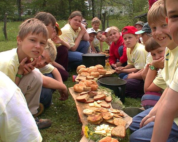 Die Kinder sitzen im Freien auf dem Rasen in zwei Reihen einander gegenüber, und zwischen ihnen liegen Brot, Käse und andere Lebensmittel auf Holzbrettern.
