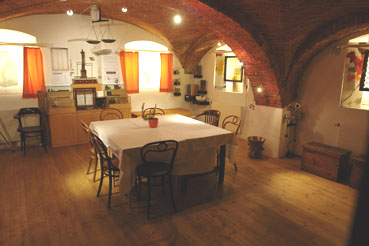 Raum mit gewölbter Ziegeldecke, Holzboden und einem Tisch mit Stühlen in der Mitte; rote Vorhänge bedecken die Fenster, und antike Möbel schmücken den Raum.