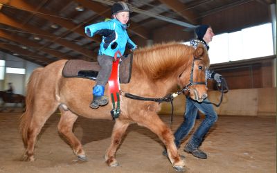 Ein kleines Kind in einer blauen Jacke reitet im Haus auf einem Pony, während ein Erwachsener das Pony an einem Seil führt.
