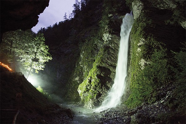 Ein Wasserfall fließt nachts einen von Bäumen umgebenen Felsen hinunter, der von der linken Seite des Bildes aus mit künstlichem Licht beleuchtet wird.