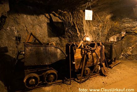 In einer schwach beleuchteten unterirdischen Höhle mit rauen Felswänden sind verrostete Bergbauwagen und -geräte ausgestellt.