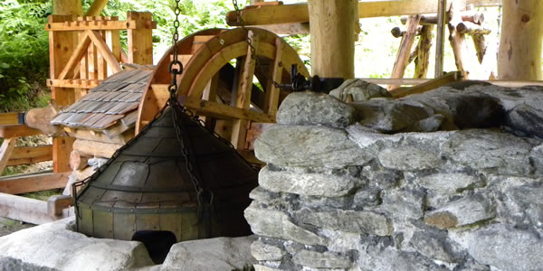 Ein traditionelles wasserbetriebenes Blasebalgsystem aus Holz und Stein, das für die Metallbearbeitung verwendet wird, ist im Freien unter einem hölzernen Unterstand aufgestellt.