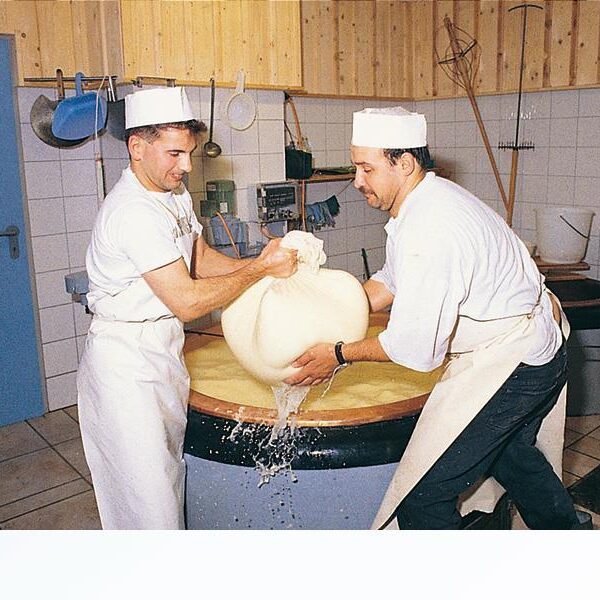 Zwei Männer in weißen Uniformen und mit Hüten heben ein großes Tuch, das mit Käsebruch gefüllt ist, aus einem Bottich mit Flüssigkeit in einem Molkerei-Verarbeitungsraum.