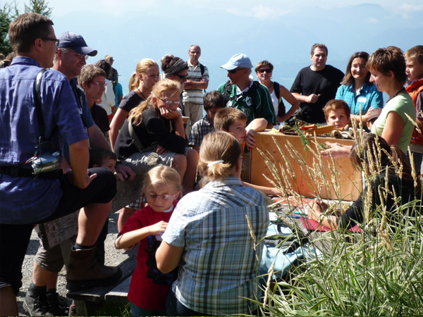 Eine Gruppe von Menschen, darunter Kinder und Erwachsene, versammelt sich im Freien um eine Frau, die einen hölzernen Gegenstand hält; im Hintergrund sind Berge zu sehen.