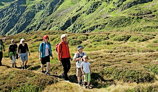 Eine Gruppe von Menschen, darunter Kinder und Erwachsene, wandert an einem sonnigen Tag auf einem grasbewachsenen Bergpfad, umgeben von grünen Hügeln.