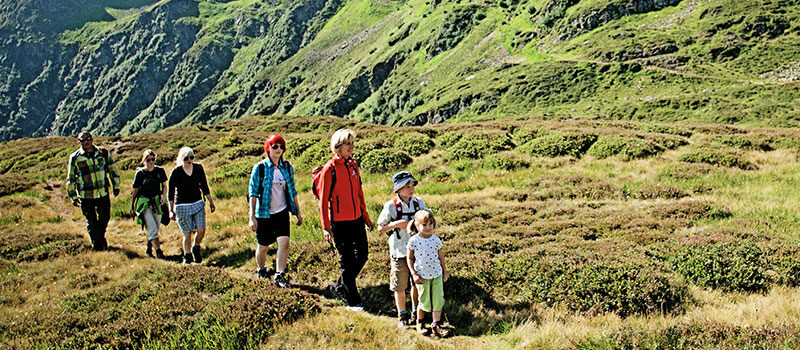 Eine Gruppe von Menschen, darunter Kinder und Erwachsene, wandert an einem sonnigen Tag auf einem grasbewachsenen Bergpfad, umgeben von grünen Hügeln.