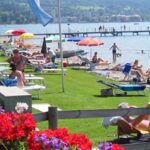 Menschen, die sich auf Liegestühlen sonnen und unter Sonnenschirmen auf einer Wiese und am Sandstrand eines Sees entspannen, mit Bergen im Hintergrund.
