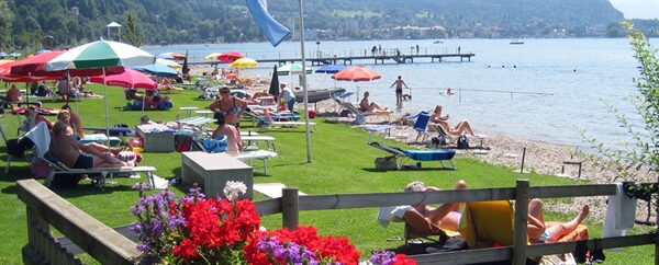 Menschen, die sich auf Liegestühlen sonnen und unter Sonnenschirmen auf einer Wiese und am Sandstrand eines Sees entspannen, mit Bergen im Hintergrund.