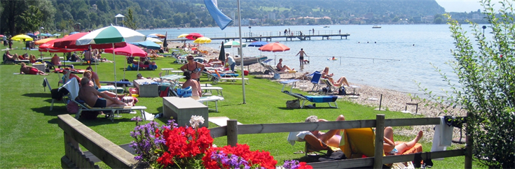 Menschen, die sich auf Liegestühlen sonnen und unter Sonnenschirmen auf einer Wiese und am Sandstrand eines Sees entspannen, mit Bergen im Hintergrund.