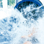 Eine Person verlässt eine Wasserrutsche in einem Innenraum, wobei am Ende der Rutsche ein großer Wasserspritzer entsteht.