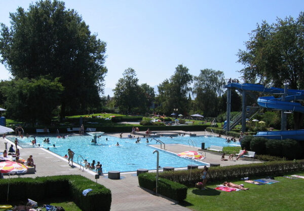 An einem sonnigen Tag schwimmen und entspannen sich die Menschen in einem Freibad mit blauer Wasserrutsche, umgeben von Bäumen und Rasenflächen.