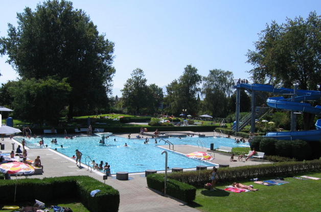 An einem sonnigen Tag schwimmen und entspannen sich die Menschen in einem Freibad mit blauer Wasserrutsche, umgeben von Bäumen und Rasenflächen.