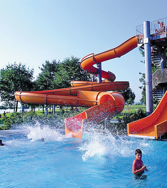 Eine orangefarbene Wasserrutsche führt in ein Schwimmbecken, in dem Kinder an einem sonnigen Tag im Wasser planschen. Im Hintergrund sind Bäume und Gras zu sehen.