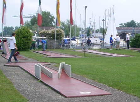 Menschen spielen Minigolf auf einem Außenplatz in der Nähe eines Jachthafens mit Segelbooten und Fahnenmasten im Hintergrund.