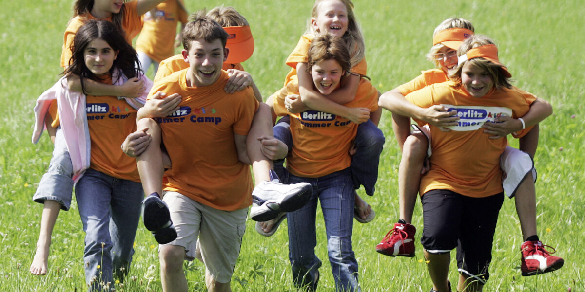 Eine Gruppe von Kindern, die orangefarbene Berlitz Sommercamp-Shirts tragen, rennen und fahren Huckepack auf einer Wiese.