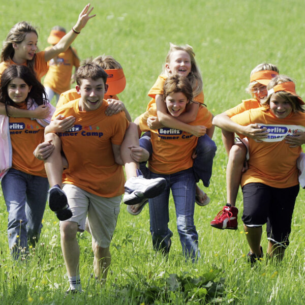 Eine Gruppe von Kindern, die orangefarbene Berlitz Sommercamp-Shirts tragen, rennen und fahren Huckepack auf einer Wiese.
