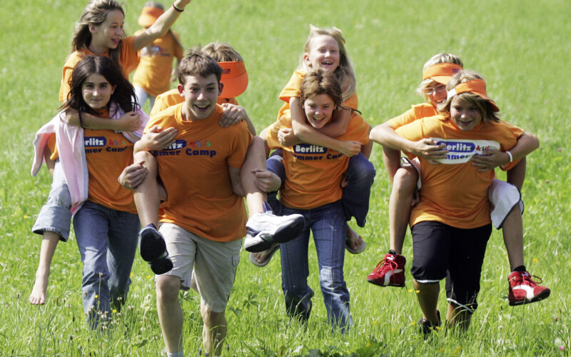 Eine Gruppe von Kindern, die orangefarbene Berlitz Sommercamp-Shirts tragen, rennen und fahren Huckepack auf einer Wiese.