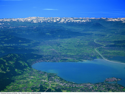 Luftaufnahme eines großen Sees, umgeben von grünen Feldern und Städten, mit entfernten Bergen unter einem klaren blauen Himmel.