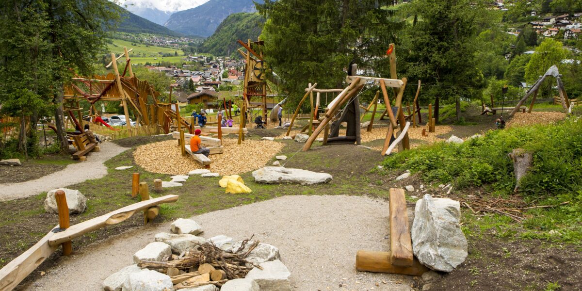 Kinderspielplatz mit Holzkonstruktionen, Klettermöglichkeiten und Bänken, eingebettet in eine bergige, grüne Landschaft mit Bäumen und entfernten Gebäuden.
