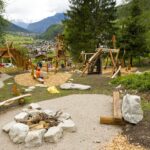 Kinderspielplatz mit Holzkonstruktionen, Klettermöglichkeiten und Bänken, eingebettet in eine bergige, grüne Landschaft mit Bäumen und entfernten Gebäuden.