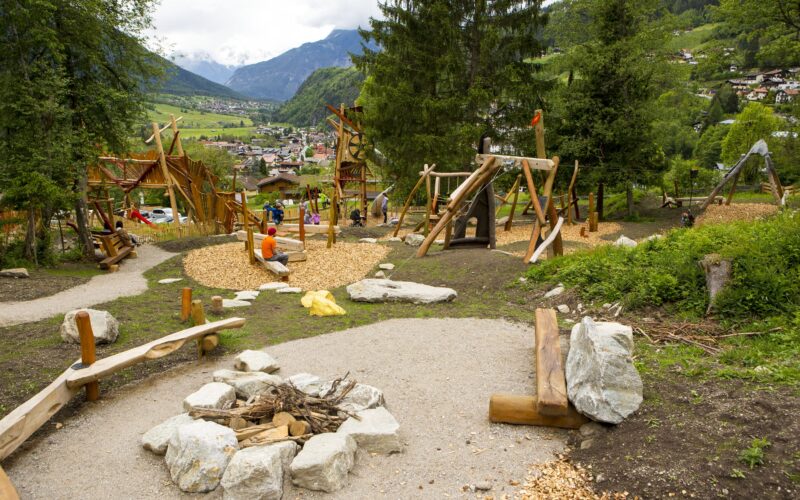 Kinderspielplatz mit Holzkonstruktionen, Klettermöglichkeiten und Bänken, eingebettet in eine bergige, grüne Landschaft mit Bäumen und entfernten Gebäuden.