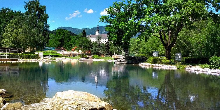 In einem ruhigen, von Felsen und Bäumen umgebenen Teich spiegelt sich ein entferntes weißes Gebäude mit einem Turm unter einem klaren blauen Himmel.