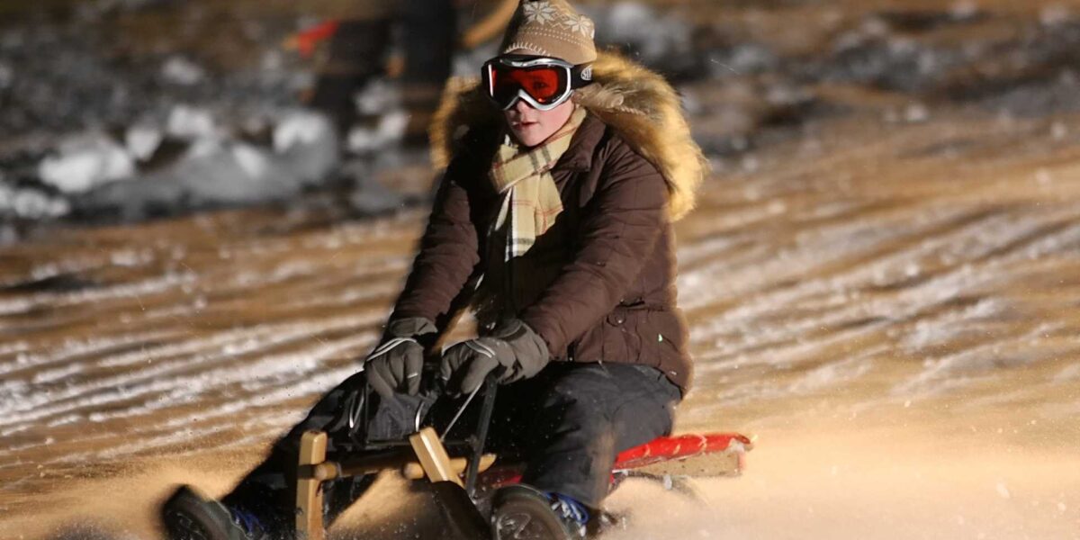 Eine Person in Winterkleidung und mit Schutzbrille fährt nachts auf einem Holzschlitten einen verschneiten Hang hinunter, wobei der Schnee um sie herum aufspritzt.
