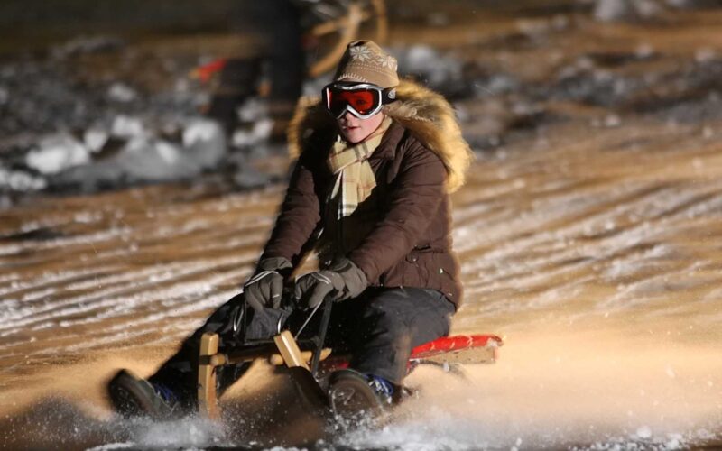 Eine Person in Winterkleidung und mit Schutzbrille fährt nachts auf einem Holzschlitten einen verschneiten Hang hinunter, wobei der Schnee um sie herum aufspritzt.