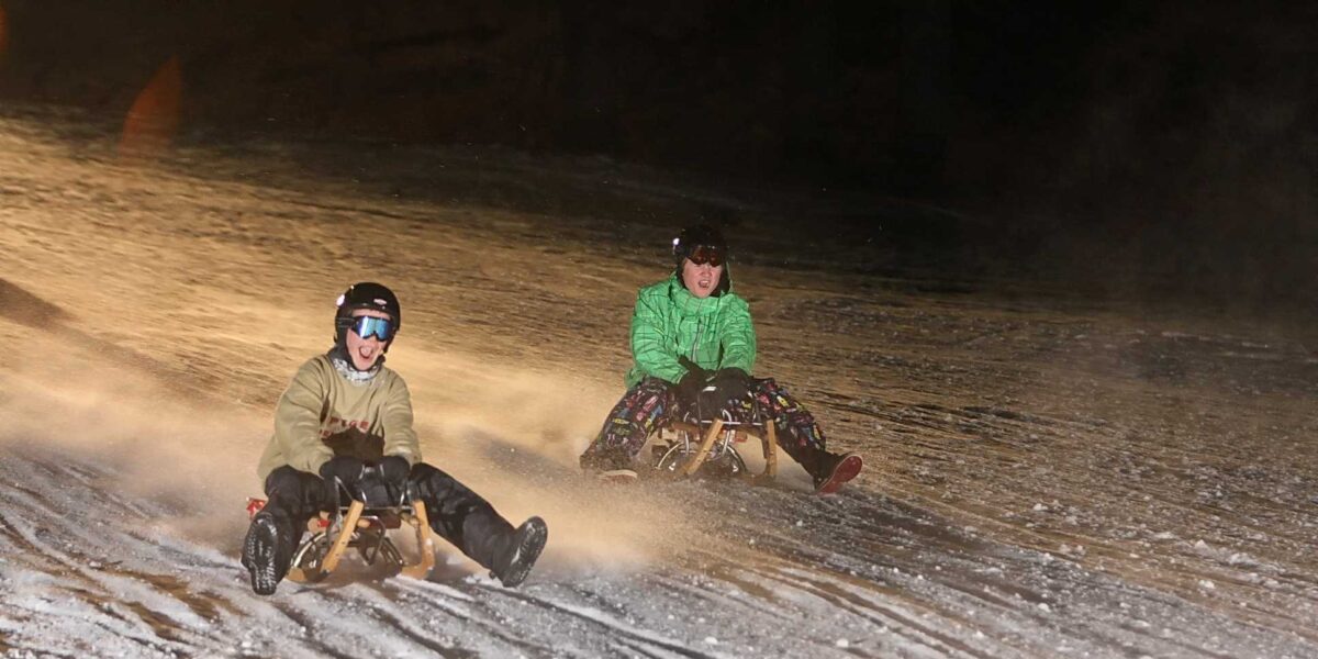 Zwei Personen mit Helmen und Winterjacken fahren nachts auf Holzschlitten einen verschneiten Hügel hinunter.