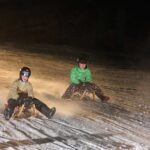 Zwei Personen mit Helmen und Winterjacken fahren nachts auf Holzschlitten einen verschneiten Hügel hinunter.