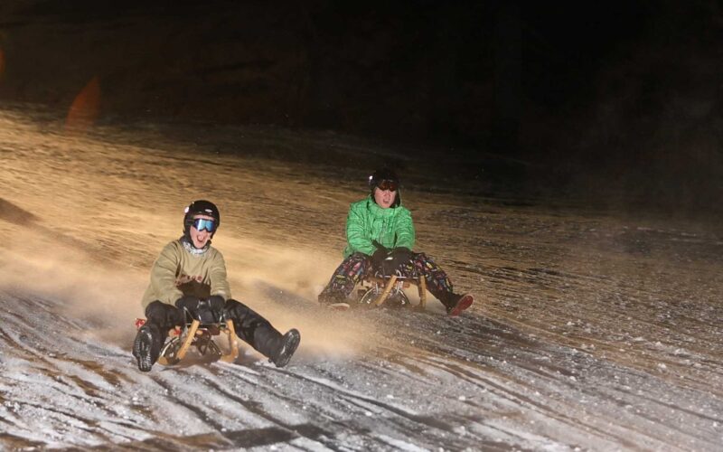 Zwei Personen mit Helmen und Winterjacken fahren nachts auf Holzschlitten einen verschneiten Hügel hinunter.