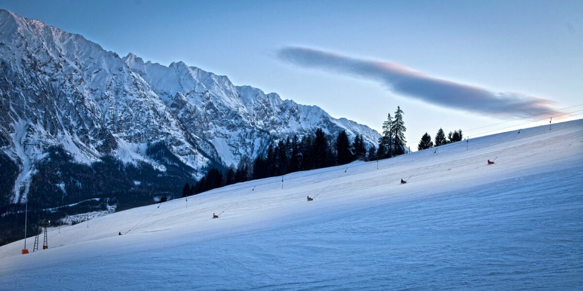 Eine schneebedeckte Skipiste, auf der mehrere Personen rodeln, umrahmt von Bäumen und einer Bergkette im Hintergrund, unter einem klaren Himmel.