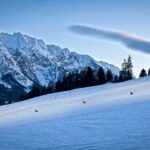 Eine schneebedeckte Skipiste, auf der mehrere Personen rodeln, umrahmt von Bäumen und einer Bergkette im Hintergrund, unter einem klaren Himmel.