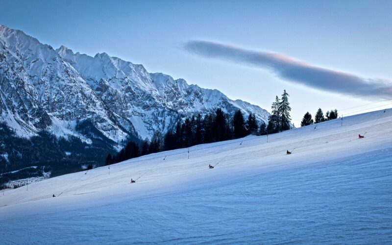 Eine schneebedeckte Skipiste, auf der mehrere Personen rodeln, umrahmt von Bäumen und einer Bergkette im Hintergrund, unter einem klaren Himmel.