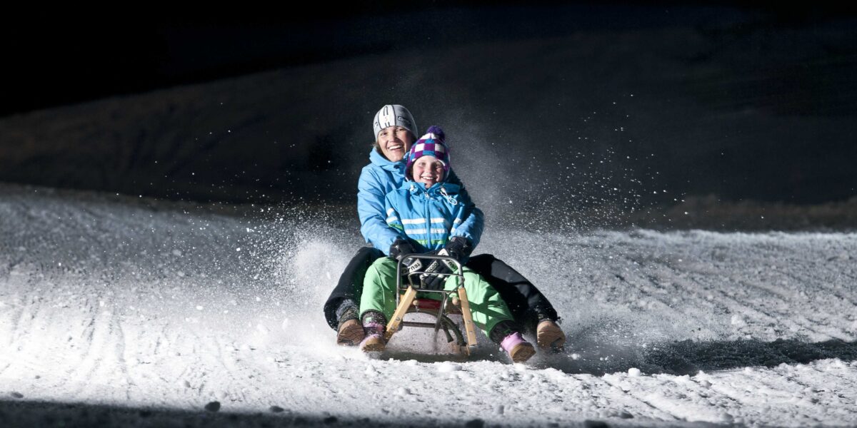 Zwei winterlich gekleidete Personen fahren nachts auf einem Holzschlitten über den verschneiten Boden, während der Schnee hinter ihnen aufspritzt.