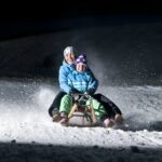 Zwei winterlich gekleidete Personen fahren nachts auf einem Holzschlitten über den verschneiten Boden, während der Schnee hinter ihnen aufspritzt.
