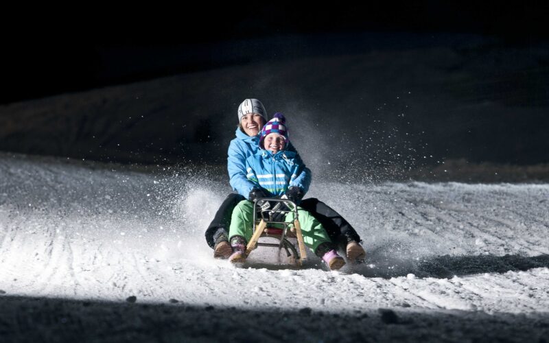 Zwei winterlich gekleidete Personen fahren nachts auf einem Holzschlitten über den verschneiten Boden, während der Schnee hinter ihnen aufspritzt.