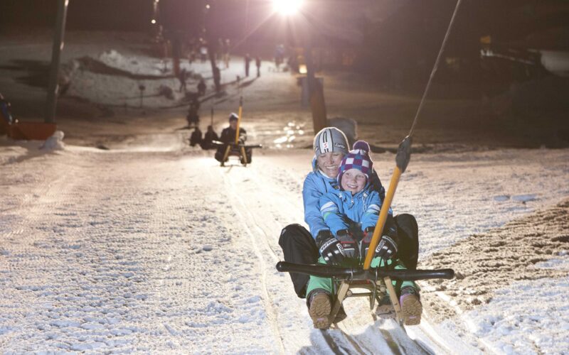 Zwei Personen fahren nachts mit einem Schlitten einen verschneiten Hang hinauf, wobei sie sich an einem Liftgriff festhalten, während im Hintergrund andere Schlittenfahrer und Lichter zu sehen sind.