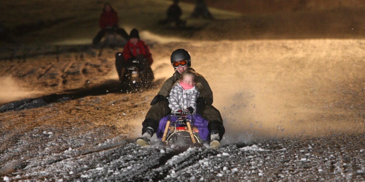 Eine Person mit Helm und Schutzbrille fährt mit einem Kind auf einem Schlitten bergab, auf einer verschneiten Strecke bei Nacht, während im Hintergrund andere Schlitten fahren.