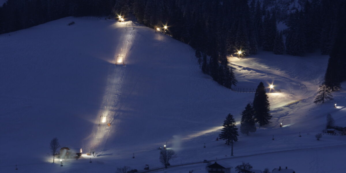 Eine schneebedeckte Skipiste wird nachts von mehreren Lichtern beleuchtet. Am Fuße und am Rande der Piste sind einige kleine Gebäude und Bäume zu sehen.