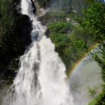 Ein hoher Wasserfall stürzt in Kaskaden einen von grünen Bäumen umgebenen Felshang hinunter, und am Fuße ist im Nebel ein Regenbogen zu sehen.