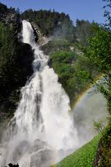 Ein hoher Wasserfall stürzt in Kaskaden einen von grünen Bäumen umgebenen Felshang hinunter, und am Fuße ist im Nebel ein Regenbogen zu sehen.
