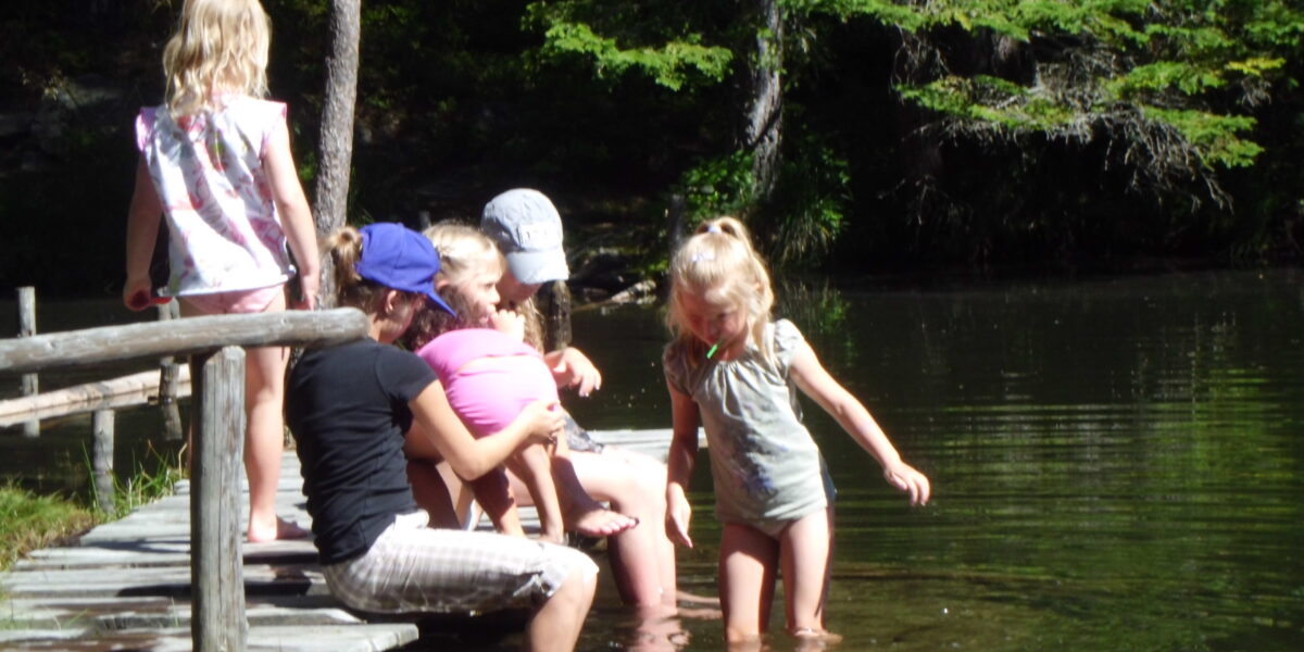 Fünf Kinder sitzen und spielen auf einem hölzernen Steg an einem bewaldeten See. Zwei von ihnen stehen im seichten Wasser, die anderen sitzen oder knien auf dem Steg im Sonnenlicht.