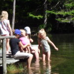 Fünf Kinder sitzen und spielen auf einem hölzernen Steg an einem bewaldeten See. Zwei von ihnen stehen im seichten Wasser, die anderen sitzen oder knien auf dem Steg im Sonnenlicht.