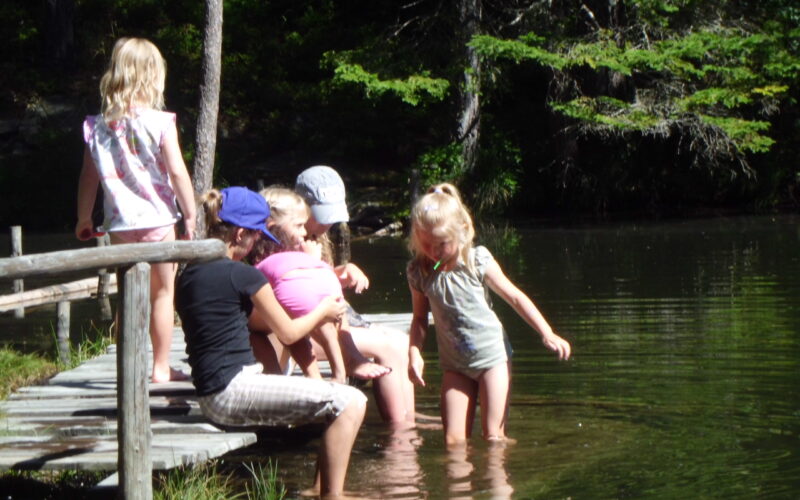 Fünf Kinder sitzen und spielen auf einem hölzernen Steg an einem bewaldeten See. Zwei von ihnen stehen im seichten Wasser, die anderen sitzen oder knien auf dem Steg im Sonnenlicht.