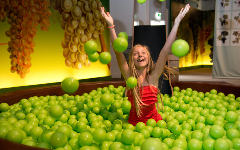 Ein Mädchen in einem roten Kleid wirft grüne Plastikbälle, während es in einer großen, mit grünen Bällen gefüllten Grube sitzt.