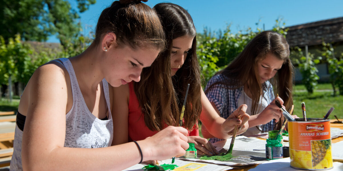 Drei junge Frauen sitzen an einem Tisch im Freien und beschäftigen sich unter sonnigem Himmel mit grünen Farben und Pinseln mit Malprojekten.